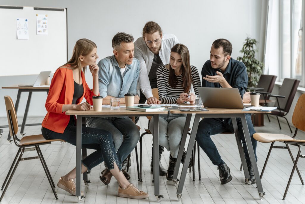 Group of colleagues collaborating at a table in an office, reviewing documents and discussing work while using a laptop.