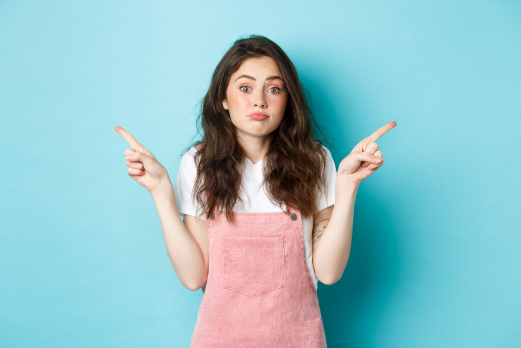 Young woman shrugging and pointing in opposite directions with a confused expression against a blue background.