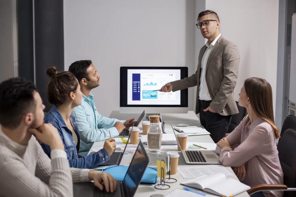 Business professional presenting data on a screen to a group of colleagues in a meeting, while participants listen and work on laptops around a conference table.