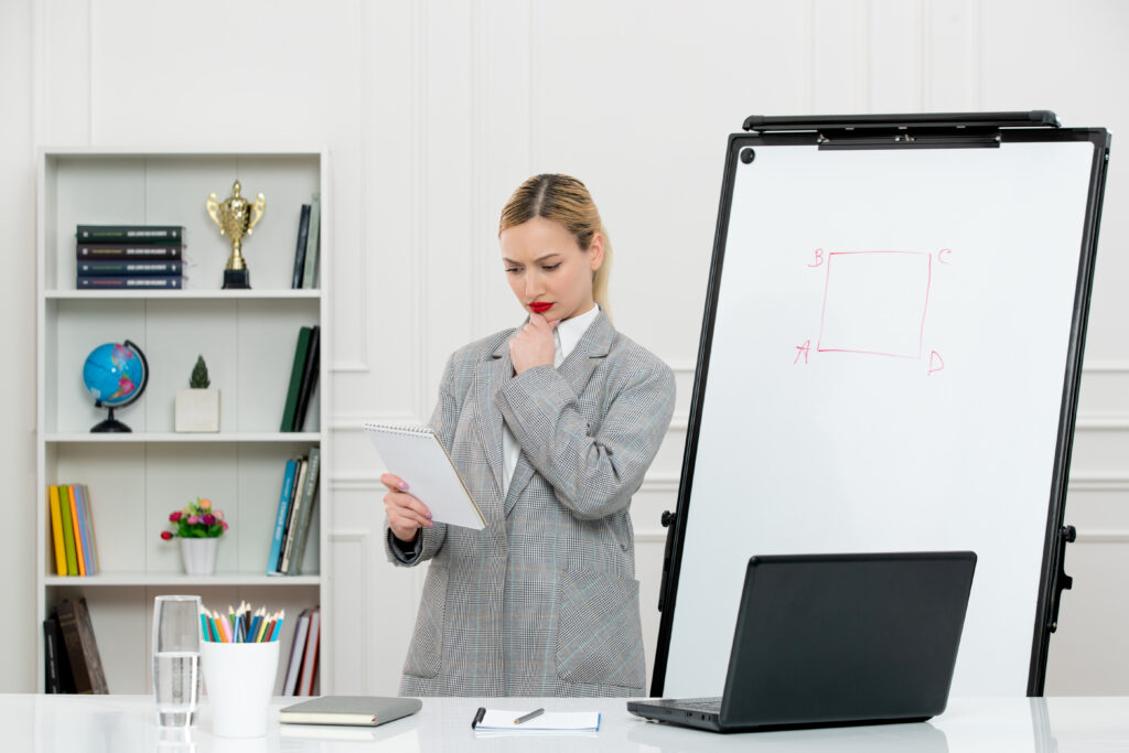 Teacher standing beside a whiteboard with a drawn diagram, holding a notebook and thoughtfully reviewing notes in a classroom or office setting.