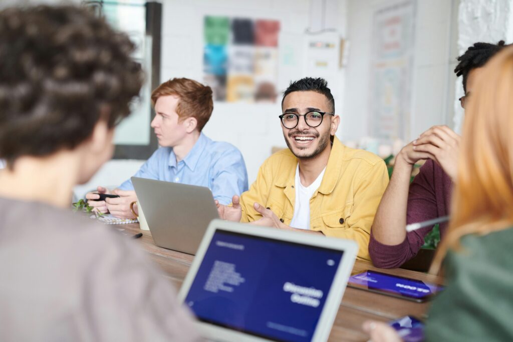 A man in a yellow shirt and glasses is smiling over at another person. They're both using laptops.