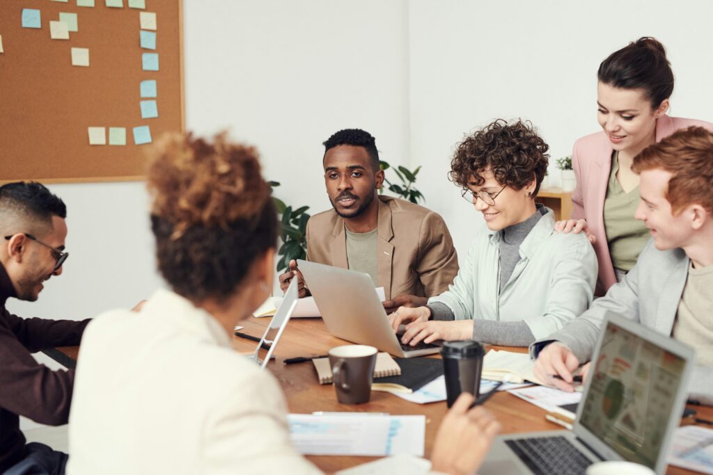 One person with curly hair and glasses is typing on a laptop and smiling. There are five other people also doing work. Two are on laptops, one has some notes and the fifth is standing and smiling.
