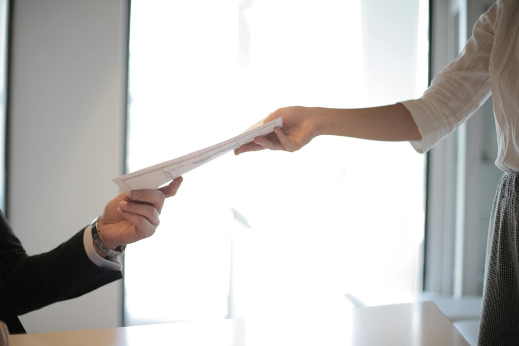 A woman giving a man a piece of paper. Their faces aren't in shot but you can tell the man is sitting down and the woman is standing.