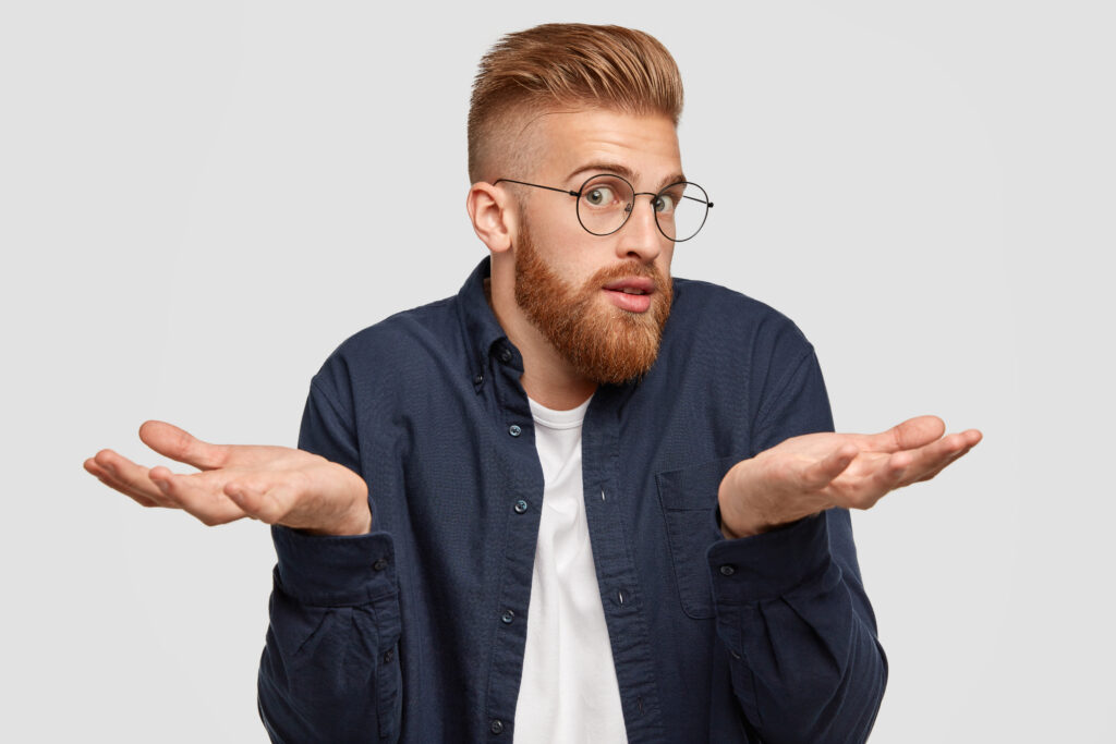 Photo of confused bearded ginger male in spectacles looks surprisingly at camera, shruggs shoulders with bewilderment, hesitates about something, has doubtful expression, isolated on white wall
