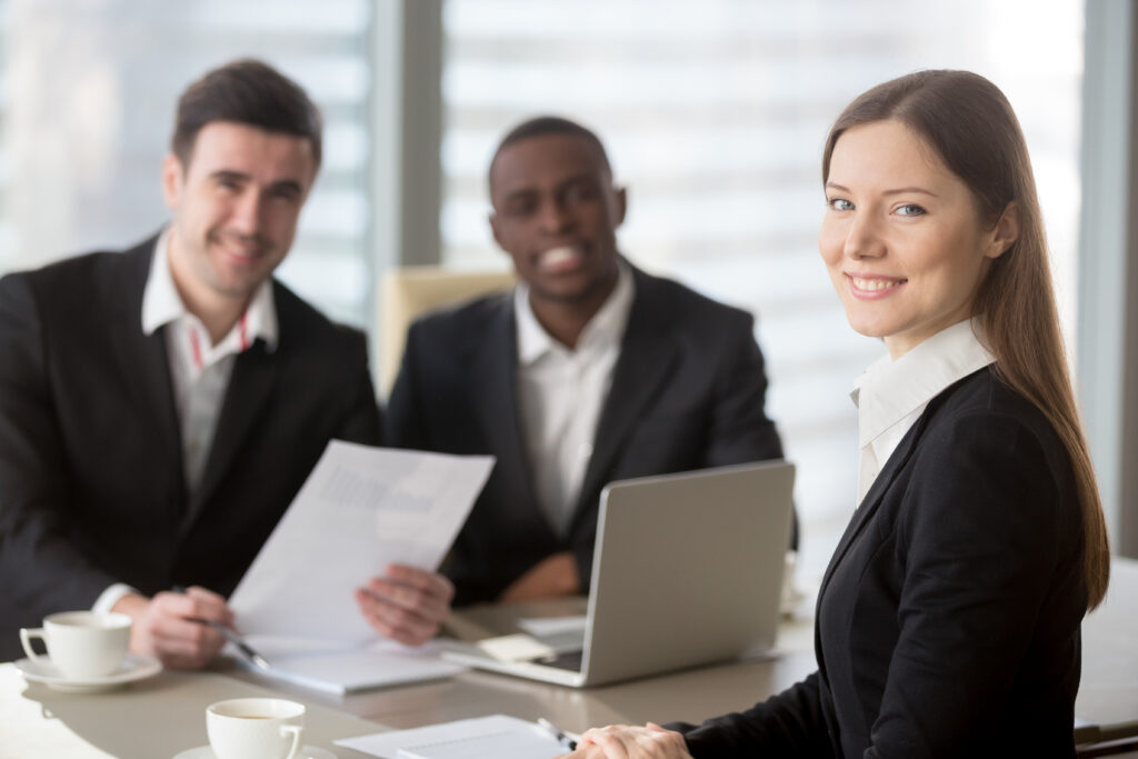 Attractive young businesswoman smiling for camera sitting at desk, young executive manager holding business meeting for black and white subordinates clients at background, female team lead portrait