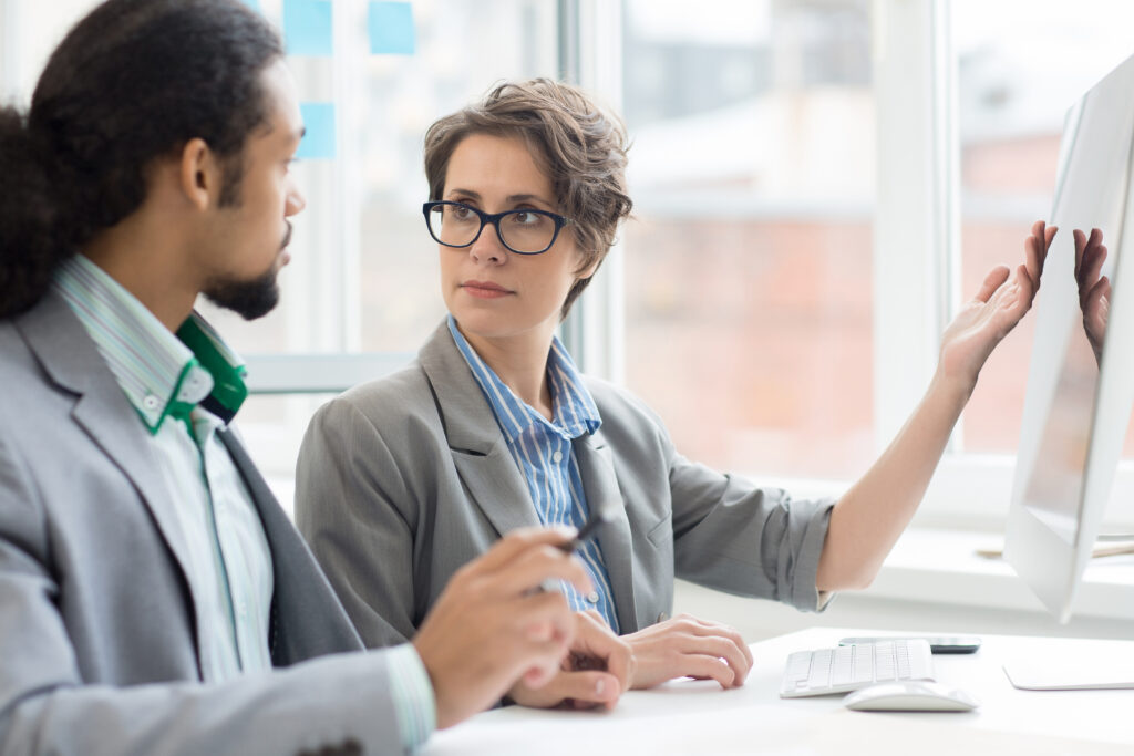 Serious female trader pointing at computer monitor and looking at her colleague during discussion of online financial statistics